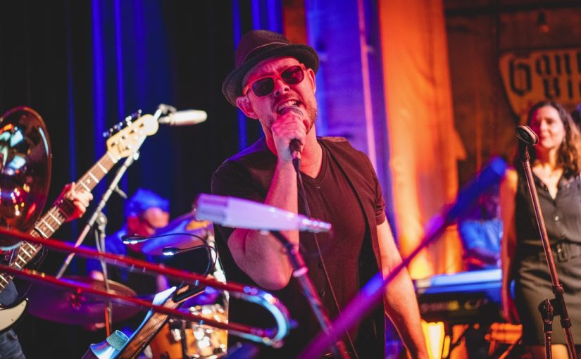 Mehrere Musiker der Band Soulfood Café stehen auf einer Bühne mit bunten Bühnenlichtern in Blau und Orange. Im Vordergrund hält ein Sänger ein Mikrofon und trägt ein schwarzes T-Shirt sowie einen Hut. Links ist ein Bassgitarrist mit einem hellen Bass zu sehen, im Hintergrund ein Keyboardspieler. Rechts steht eine Sängerin mit Mikrofonständer. Die Bühne ist mit verschiedenen Instrumenten und Mikrofonen ausgestattet, die Atmosphäre wirkt lebendig und musikalisch.