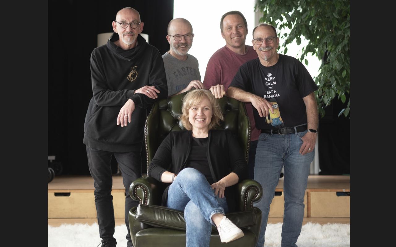 Eine Gruppe von fünf fröhlichen Personen, die Band Pretty Acoustic, ist in einem Innenraum mit dunklem Hintergrund und einer großen grünen Pflanze rechts zu sehen. Eine Frau mit blonden Haaren sitzt entspannt in einem dunkelgrünen Ledersessel mit hohen Armlehnen, trägt eine schwarze Jacke, blaue Jeans und weiße Sneaker. Vier Männer, drei bärtige mit Brille, stehen hinter dem Sessel. Die Person ganz links trägt einen schwarzen Kapuzenpullover mit einem goldfarbenen Emblem auf der Brust und schwarze Hosen. Der zweite Mann von links trägt ein graues T-Shirt mit einem kaum lesbaren Aufdruck. Der dritte Mann von links trägt ein rotes T-Shirt. Der Mann ganz rechts trägt ein schwarzes T-Shirt mit einem Minion-Motiv und dem Text 'KEEP CALM AND EAT A BANANA' sowie eine Armbanduhr und blaue Jeans. Der Boden ist mit einem weißen Teppich bedeckt, und im Hintergrund sind Holzbühnenstufen zu erkennen.