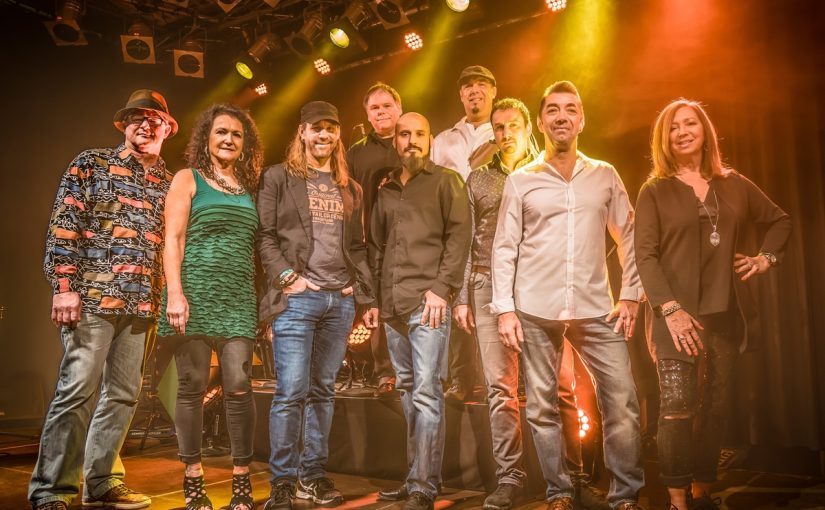 Zehn Personen der Band Fonkzone stehen auf einer Bühne vor orange-gelber Bühnenbeleuchtung, einige tragen legere Kleidung wie Jeans und Hemden, eine Person trägt ein grünes Kleid.